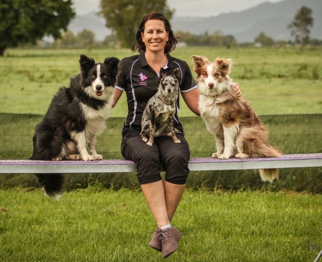 Woman with dark hair hair smiling and facing the camera.  She is sitting on a plank with a small dog on her lap and a border collie on either side of her.