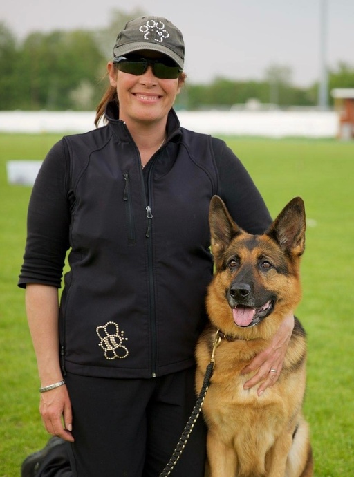 Woman wearing ball cap sitting in grass field smiling, facing camera with her arm around a german shepherd