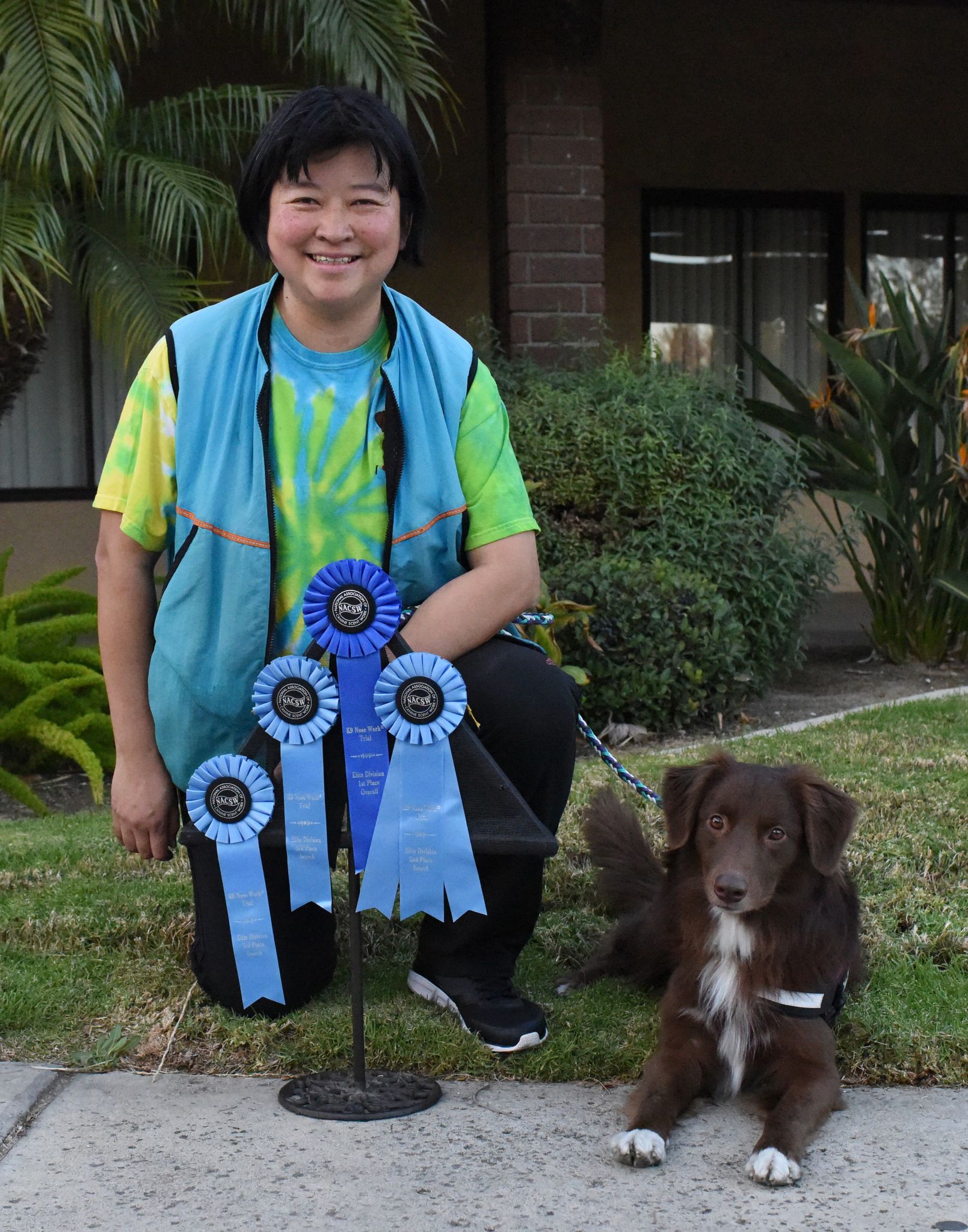 Woman with dark hair smiling with a brown australian shepherd holding 4 blue ribbons with a building and trees in the background