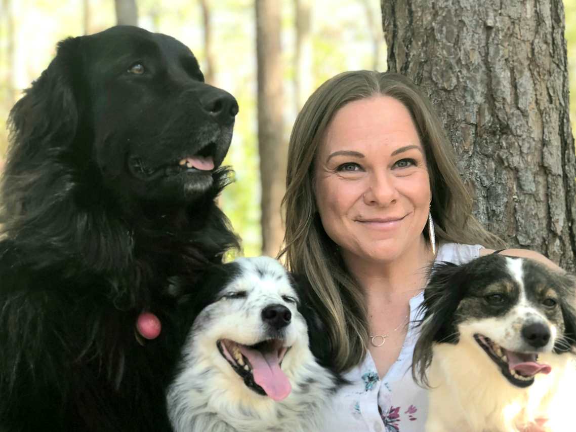Woman with shoulder length brown hair smiling and facing the camera.  She is surrounded by three dogs.