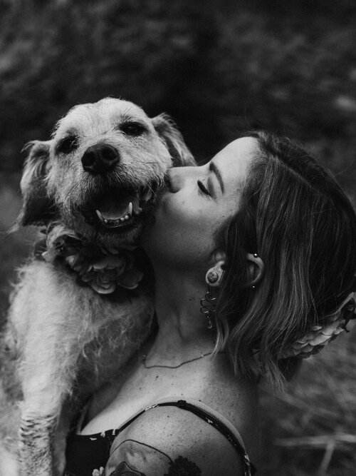 Woman with shoulder length hair smiling and kissing her dog. The dog is smiling facing the camera.  Picture is in black and white.