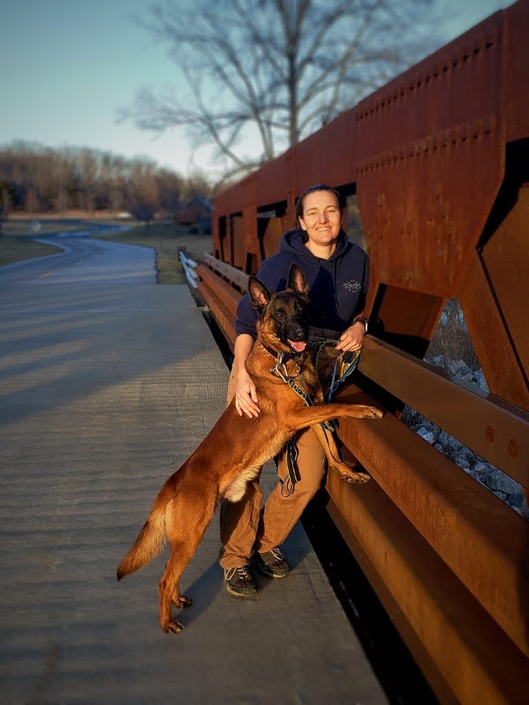 Woman with pulled back dark hair hair smiling and facing the camera.  She is leaning against a metal bridge and has a malinois dog posing with her.