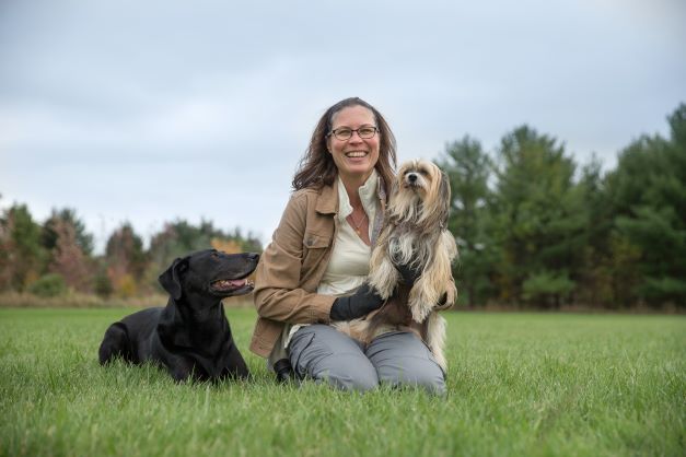 Woman with shoulder length dark hair sitting in grass field smiling, facing camera holding a chinese crested dog with a black lab cross sitting beside her