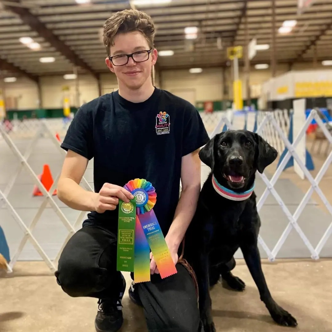 Man with short brown hair and glasses posing with black labrador retriever holding ribbons in front of a ring