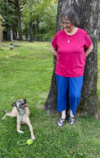 Woman with dark hair leaning against a tree smiling down at a blond dog that is smiling up at her