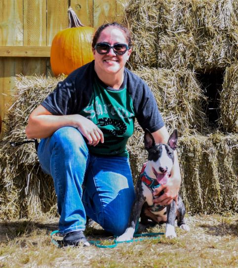 Woman with dark hair and sunglasses smiling facing camera crouched down next to a black and white bull terrier