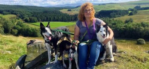 Woman with shoulder length curly red hair smiling and facing the camera.  She is surrounded by three dogs with fields and trees in the background.