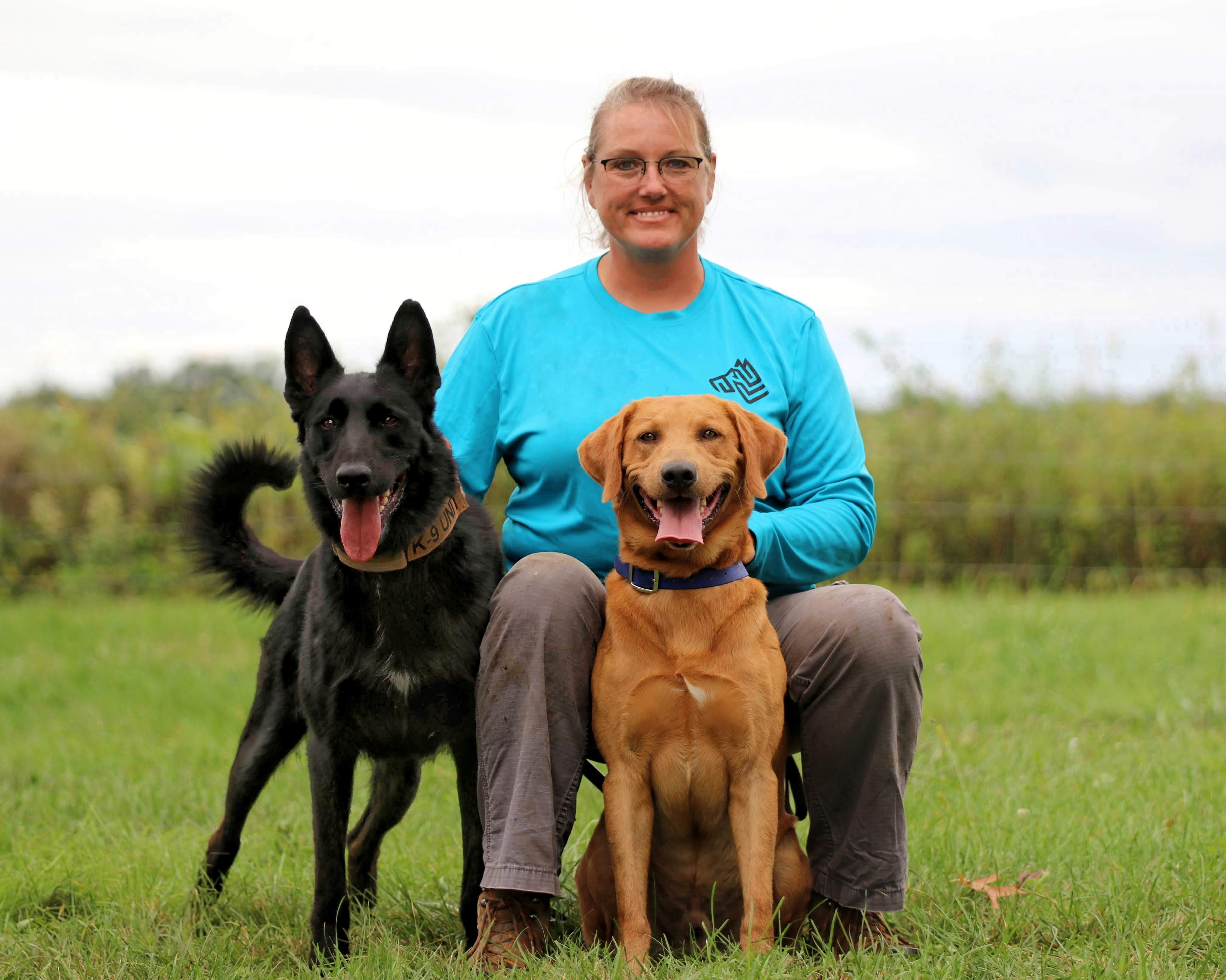 Woman with blond hair pulled back wearing a blue shirt sitting in a field with a labrador and a shepherd dog