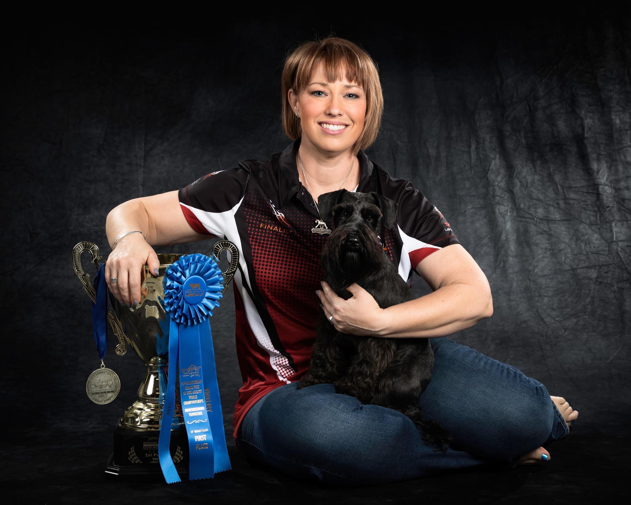 Woman with chin length brown hair smiling towards camera holding a black mini schnauzer and leaning on a large trophy, blue ribbon and medal