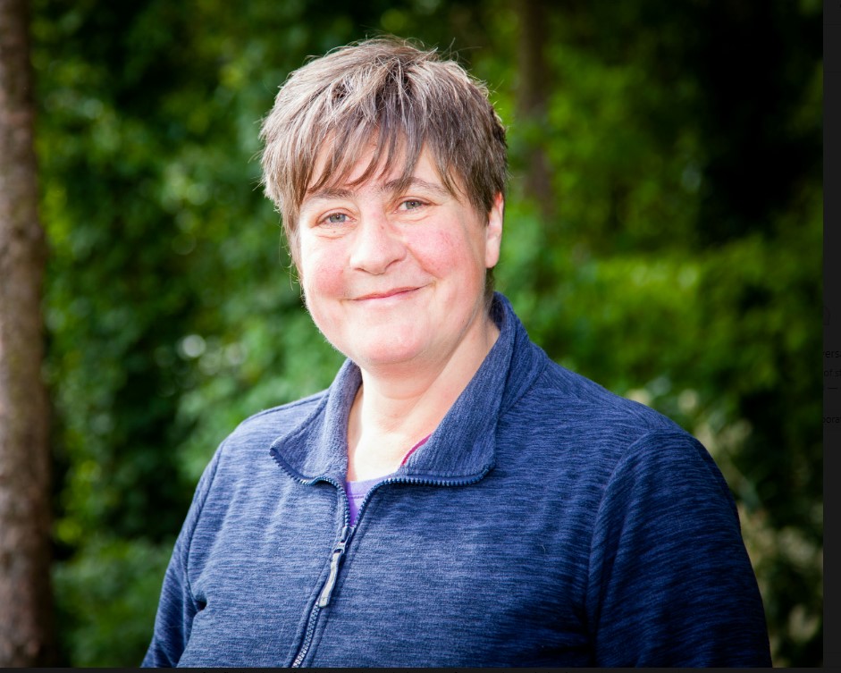 Woman with short brown hair smiling and facing slightly away from the camera with green trees in the background